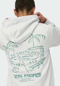 Haut de hoodie léger en gris clair, fabriqué en tissu doux, avec un design graphique vert représentant des palmier, une planche de surf et du texte au dos. La capuche est ajustable.