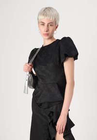 Juoda peplum suknelė su pūstu rankovėmis, tekstūruotu raštu ir raukinėtu apvadu. Modelis laiko mažą juodą rankinę su metaline juostele.