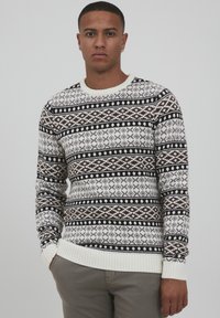 Pull en tricot avec une base crème et des motifs géométriques marron et noir. Tissu texturé avec un col et des poignets côtelés.