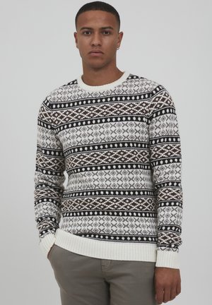 Knit sweater featuring a cream base with geometric patterns in brown and black. Textured fabric with a ribbed neckline and cuffs.