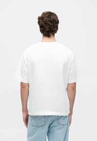 Biele bavlnené tričko s krátkymi rukávmi a rovým strihom, zobrazené zozadu, doplnené svetlomodrými džínsami.