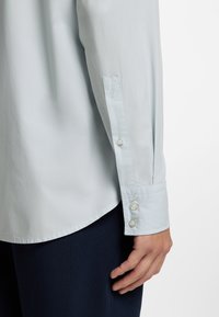 Ανοιχτό μπλε πουκάμισο με μακριά μανίκια, που διαθέτει μανσέτα με τρία μαργαριταρένια κουμπιά, απαλό ύφασμα και χαλαρή γραμμή.