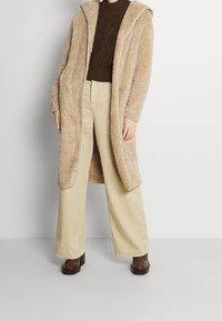 Beżowy, puszysty długi płaszcz z kapturem, w połączeniu z kremowymi spodniami o szerokich nogawkach i ciemnobrązowymi butami na grubym obcasie. Przyjemna tekstura i luźny krój.