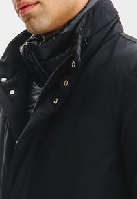 Czarna kurtka z wysokim kołnierzem, z zamkiem błyskawicznym i zatrzaskami, wykonana z gładkiego, wodoodpornego materiału z ocieplanym wnętrzem.