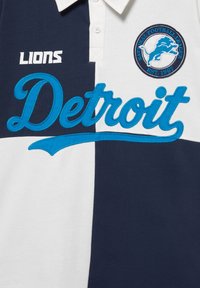 Polo avec blocs bleu marine et blanc, inscription bleue « Detroit », texte « Lions » et écusson rond avec un lion bleu et « National Football League EST. 1967. »