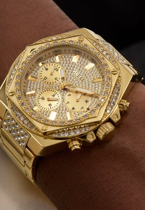 Gold-toned octagonal watch with crystal accents, featuring three subdials and a textured bracelet. Dial displays numbers and markers prominently.