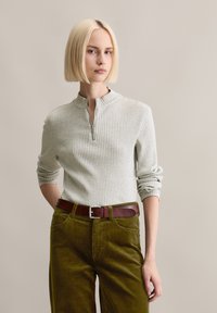 Hellgrauer gerippter Pullover mit einem Halbzip-Kragen, kombiniert mit olivgrünen Cordhose und einem braunen Leder-Gürtel mit einer silbernen Schnalle.