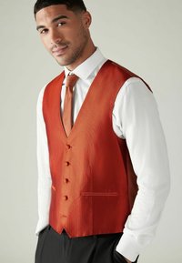 Gilet texturé orange avec revers à cran, six boutons et deux poches, associé à une chemise habillée blanche et une cravate assortie. Pantalon noir.