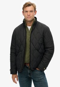 Chaquetón negro acolchado con cuello alzado, usado sobre un suéter de punto verde, combinado con jeans azules y un cinturón marrón.