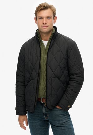 Zwarte gewatteerde jas met een staande kraag, gedragen over een groene gebreide trui, gecombineerd met blauwe jeans en een bruine riem.