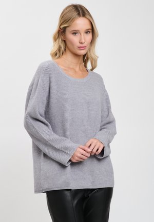 Frau mit blonden Haaren, die einen lockeren, hellgrauen, gerippten Pullover und schwarze Hosen trägt, steht vor einem schlichten Hintergrund.