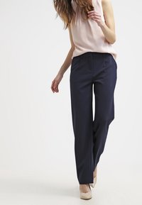 Tummansinisen väriset leveälahkeiset housut, joissa on sileä pinta, etutaskut ja vartaloa myötäilevä istuvuus, yhdistettynä vaaleanpunaisiin hihattomiin paitoihin.