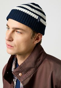 Gorro de punto a rayas en azul marino y blanco. Presenta una textura acanalada con una etiqueta de logo. Combinado con una chaqueta marrón y una camisa de cuadros.