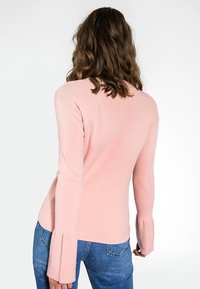 Pull long de couleur rose clair avec une coupe ajustée et des détails de manches évasées. Texture douce, porté avec un jean en denim bleu.