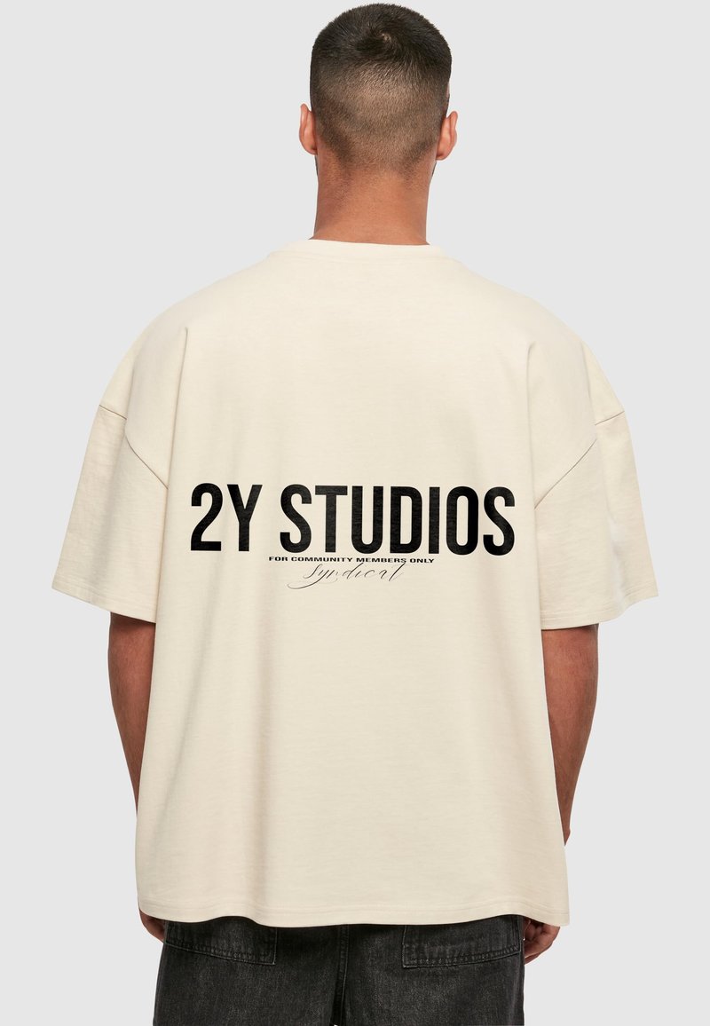 Bēša pārspīlēta T-kreklu ar melnu grafiku "2Y STUDIOS" aizmugurē, ar gludu tekstūru un īsām piedurknēm.