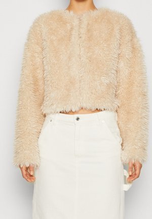 Beige fluffy cropped jakke med lange ærmer båret med højtaljet hvid nederdel, der viser maven og hænderne afslappet langs siden.