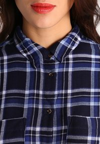 Woman wearing a blue and black plaid buttoned shirt with red lipstick, shown from chin to chest.