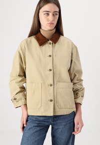 Giacca in cotone beige con colletto in velluto a coste marrone, sette bottoni, due tasche frontali e polsini con bottoni. Design casual con dettagli di cucitura visibili.
