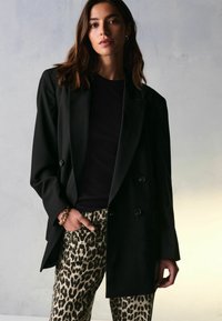 Sort dobbeltradet blazer med brede revers, parret med beige leopardmønstrede bukser. Glat stofstruktur; detalje med guldkædearmbånd.