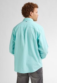 Camicia azzurro chiaro a maniche lunghe con colletto a bottoni, caratterizzata da un dettaglio sul retro e orlo arrotondato, abbinata a pantaloni grigio scuro.