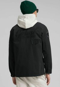 Chaqueta negra con capucha blanca, con texto en relieve y parches. Tela texturizada con puños abotonados y dobladillo recto.