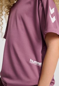 Fioletowa koszulka sportowa z teksturowanej tkaniny. Posiada białe logo "hummel" i akcenty. Krótkie rękawy z precyzyjnie wykończonymi krawędziami.