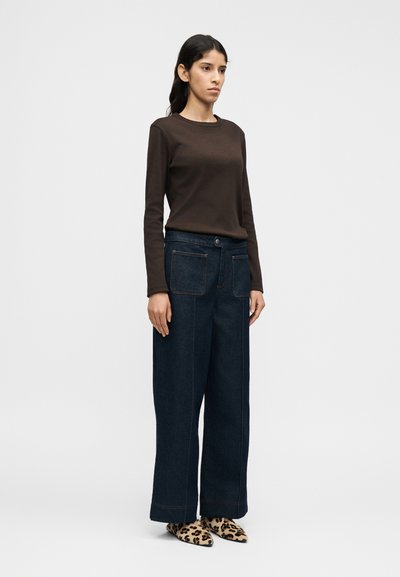 Καφέ πουκάμισο με μακριά μανίκια, σκούρο μπλε παντελόνι με φαρδιά μπότα και αντίθετη ραφή, και παπούτσια με λεοπάρ εκτύπωση και μυτερή μύτη. Απλό φόντο.