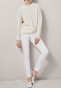 Pull en tricot à motif torsadé crème avec un col rond, associé à un jean skinny blanc et des chaussures slip-on blanches avec des accents en doré.