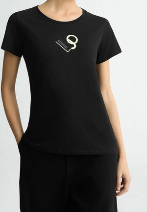 Osoba nosząca czarną krótką koszulkę z białą i czarną abstrakcyjną grafiką w pobliżu kołnierza, w połączeniu z czarnymi spodniami.