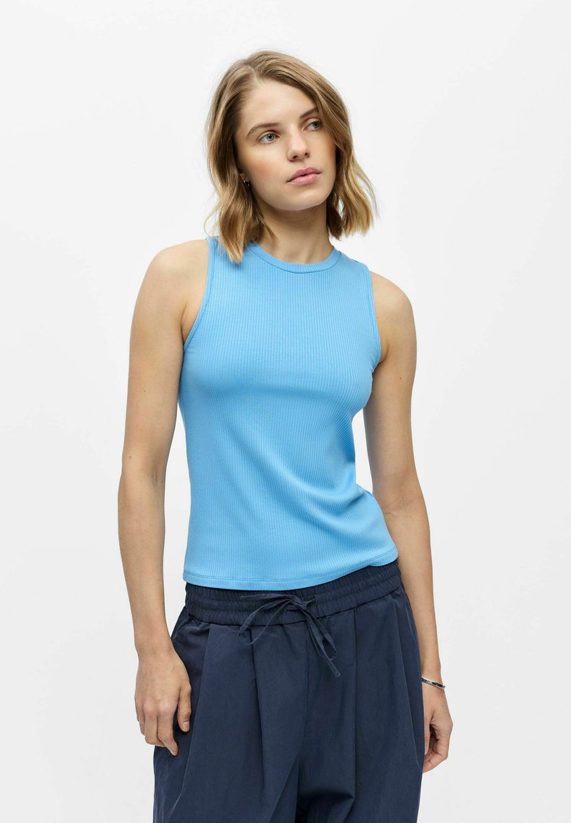 Joven mujer con cabello rubio corto que lleva una camiseta sin mangas azul claro con textura acanalada y pantalones azul oscuro con cordón, de pie frente a un fondo blanco.