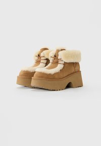 Brune semskede plattformstøvler med tykk beige saueskinnsfôr rundt ankelen og snøring, sett fra siden på hvit bakgrunn.