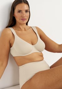 Kvinde med langt brunt hår iført beige støttende bh og højtaljede trusser, siddende afslappet mod en hvid væg.