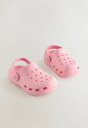 Lyserøde plastiktræsko med ventilationshuller, bagerste rem, glat tekstur og rundet tådesign. Fleksibel og let konstruktion.