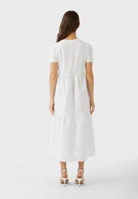 Femme aux longs cheveux bruns portant une robe midi blanche à volants avec des manches courtes et des sandales à talons hauts blanches à brides, se tenant de dos.