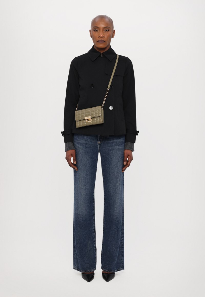 Giacca nera doppiopetto, vestibilità rilassata, abbinata a jeans blu scuri a gamba larga. Gli accessori includono una piccola borsa trapuntata verde oliva.