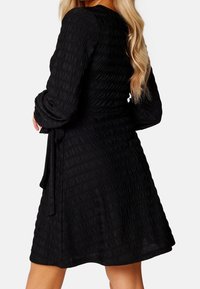 Schwarzes Kleid mit strukturiertem, gerafftem Design, langen Ärmeln und einem Bindeband in der Taille, das oberhalb der Knie endet. Glatte Stoffoberfläche.