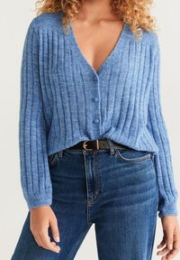 Vaaleansininen neulottu neuletakki, jossa pystysuorat resorit, V-pääntie ja kolme nappia. Yhdistetty tummansinisiin farkkuihin ja mustaan vyöhön.