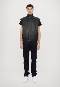 Μαύρη δερμάτινη αμάνικη γιλέκο με ψηλό γιακά, κουμπιά με πίεση και πλαϊνές τσέπες, συνδυασμένη με σκούρες παντελόνες και λευκά αθλητικά παπούτσια.