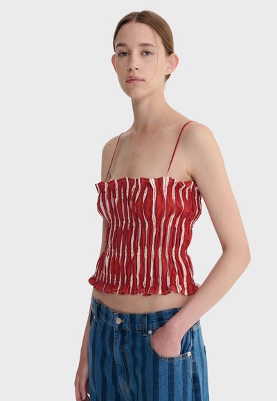 Jeune femme aux cheveux châtain clair attachés, portant un débardeur froncé à rayures rouges et blanches et un jean rayé bleu, une main dans la poche.