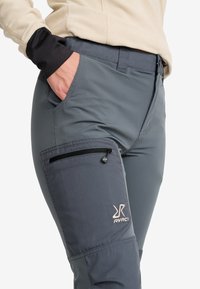 Grå, hållbara utomhusbyxor med en zippered lårficka, logodetalj och en flexibel passform. Materialet är slätt med en matt yta.