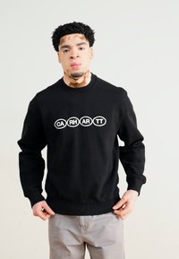Sudadera negra con cuello redondo, material de mezcla de algodón, y un logotipo circular blanco que muestra "CARHARTT" en el pecho.