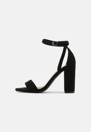 Sort højhælet sandal med ankelrem og blokhæl, åben tå og en smal rem foran.