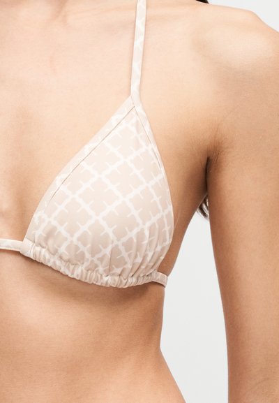 Haut de bikini beige avec motif géométrique blanc porté par une personne montrée des épaules jusqu'au milieu du torse sur fond blanc.
