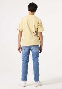 Camiseta de algodón amarilla clara con texto negro en la parte posterior, combinada con jeans de mezclilla azules y zapatillas blancas. Vista de espaldas.