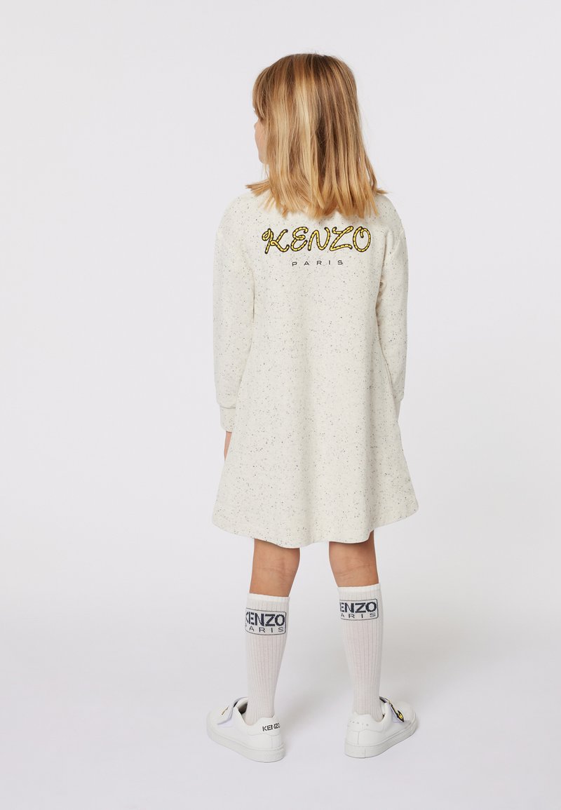 Robe blanche à pois, à manches longues, arborant le logo "KENZO PARIS" en doré au dos, associée à des chaussettes blanches jusqu'au genou et des baskets blanches.