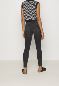 Femme portant un jean skinny noir, un haut sans manches à motifs noir et blanc, et des sandales noires, debout devant un fond blanc uni.