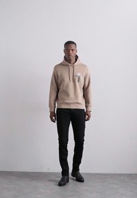 Hanorac bej cu buzunar frontal, purtat peste pantaloni negri skinny, asortat cu pantofi negri. Design simplu cu glugă și detalii cu etichetă.