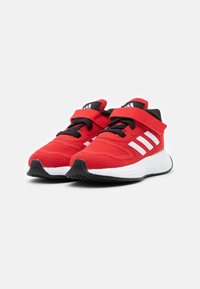 Baskets de sport rouges avec une tige en mesh, accents noirs, logo à trois bandes blanches et sangle réglable en Velcro. Semelle rembourrée pour le confort.