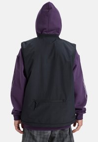 Canotta nera senza maniche sopra un hoodie viola. Presenta una tasca posteriore con zip, polsini a coste e una finitura in tessuto liscio.