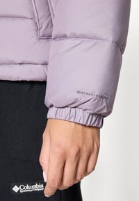 Lilla pufferjakke med elastisk mansjett, med "OMNI-HEAT" merke. Hånden hviler på et mørkt fleece-stoff.